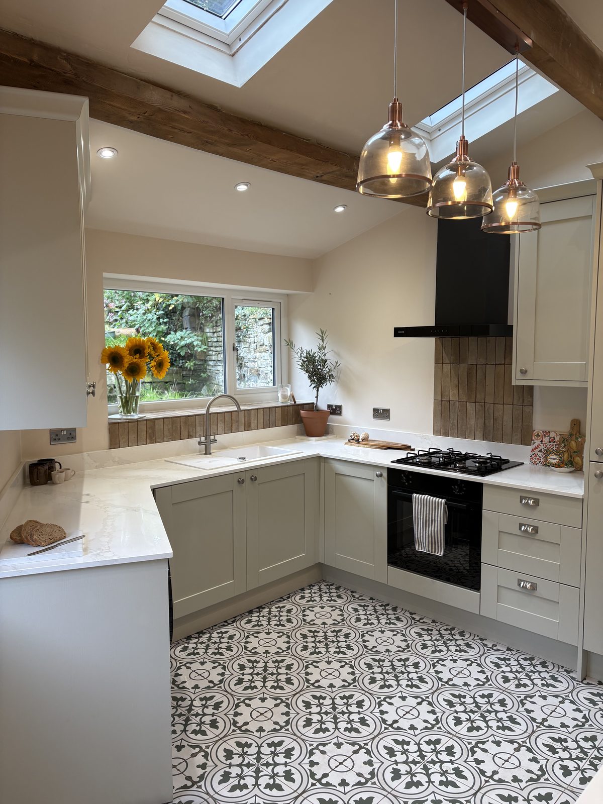 The same kitchen after renovation, with grey-green shaker cabinets, white worktops, a black extractor and patterned floor tiles